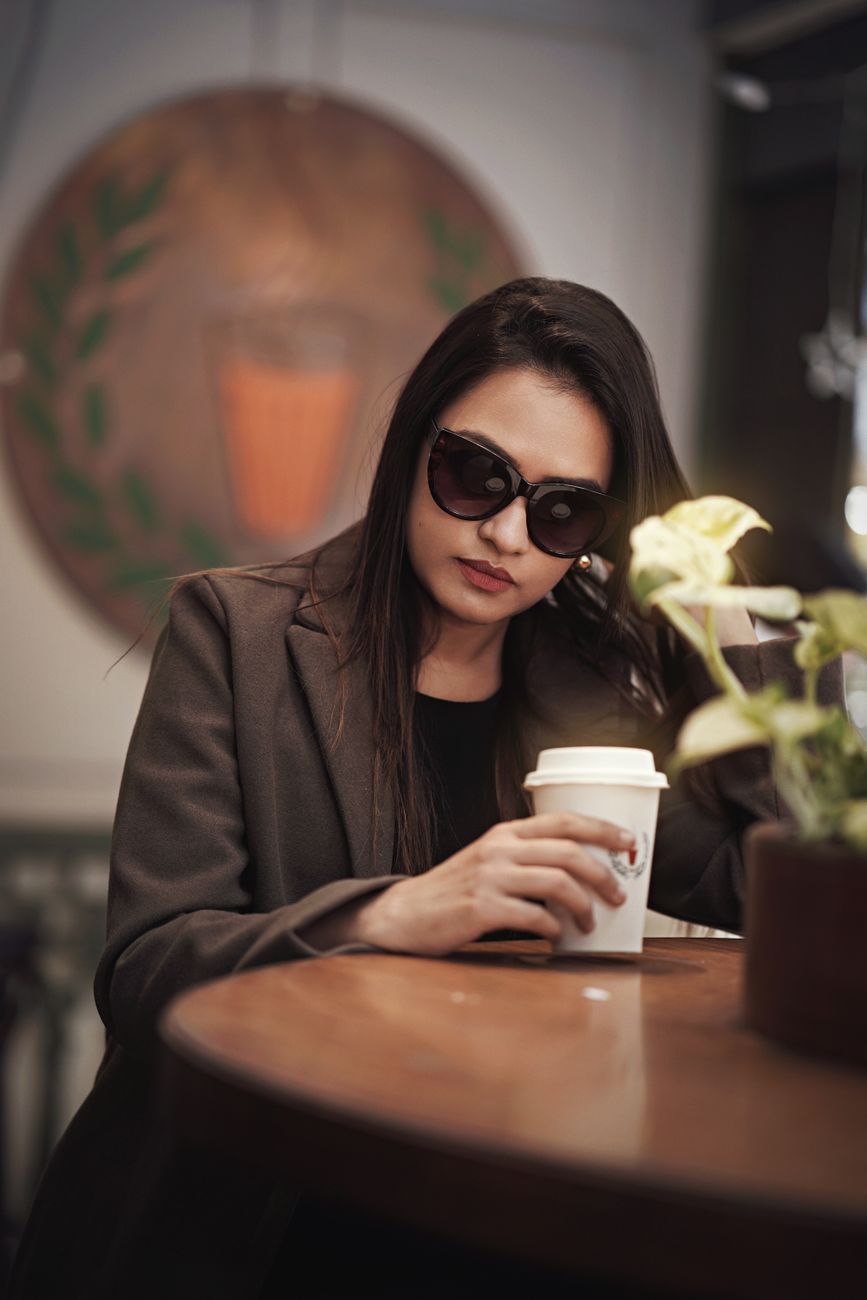 Fashionable woman in sunglasses enjoys a coffee indoors, exuding elegance and style.