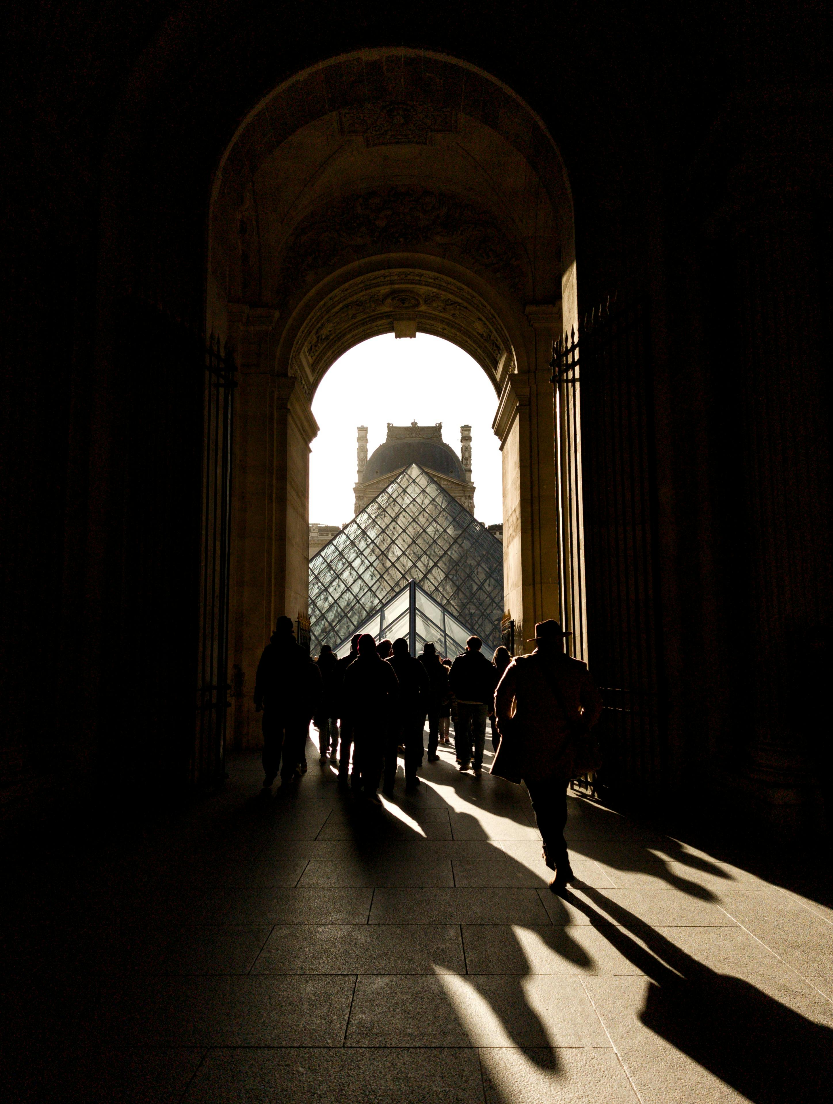 People walking through an archway towards the iconic Louvre Pyramid in Paris during daytime.