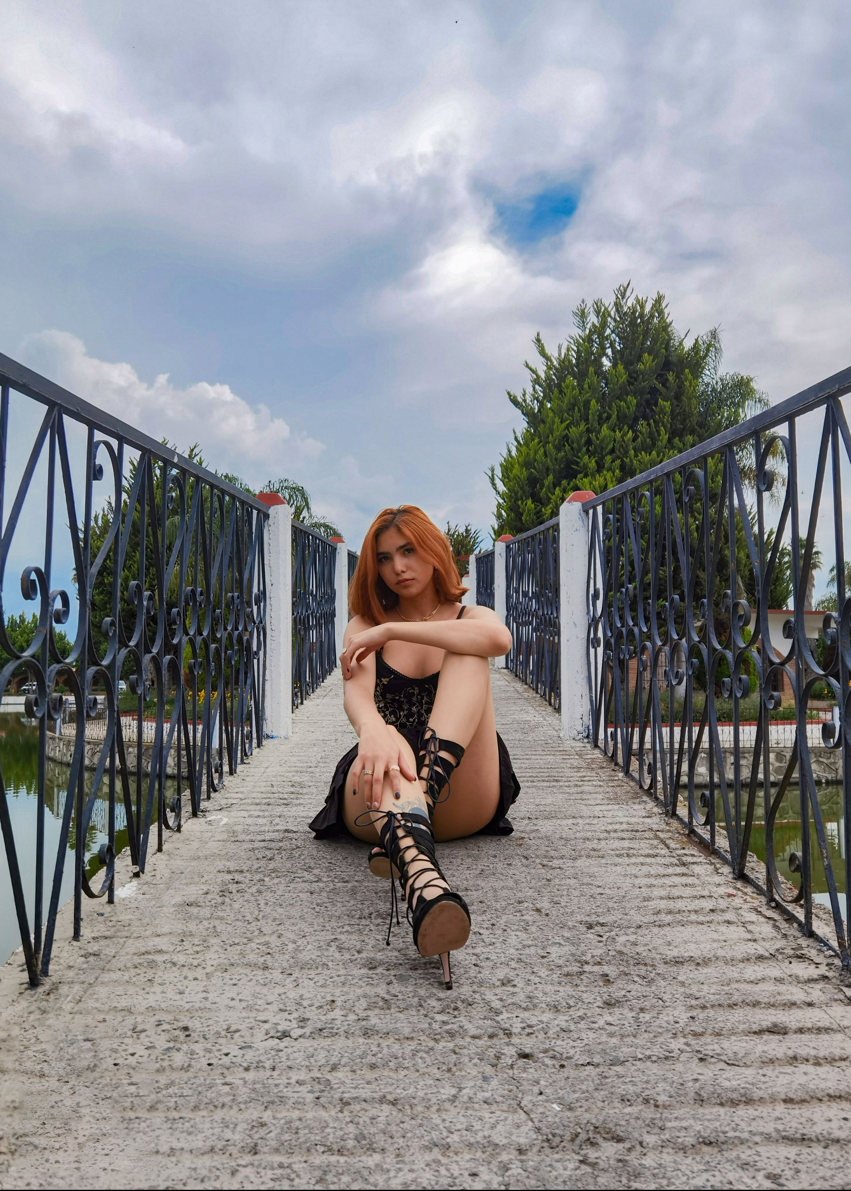 A young woman sits elegantly on a stone bridge, surrounded by lush greenery, under a cloudy sky.