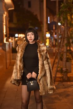 Stylish woman in fur coat holding designer bag, city street backdrop.