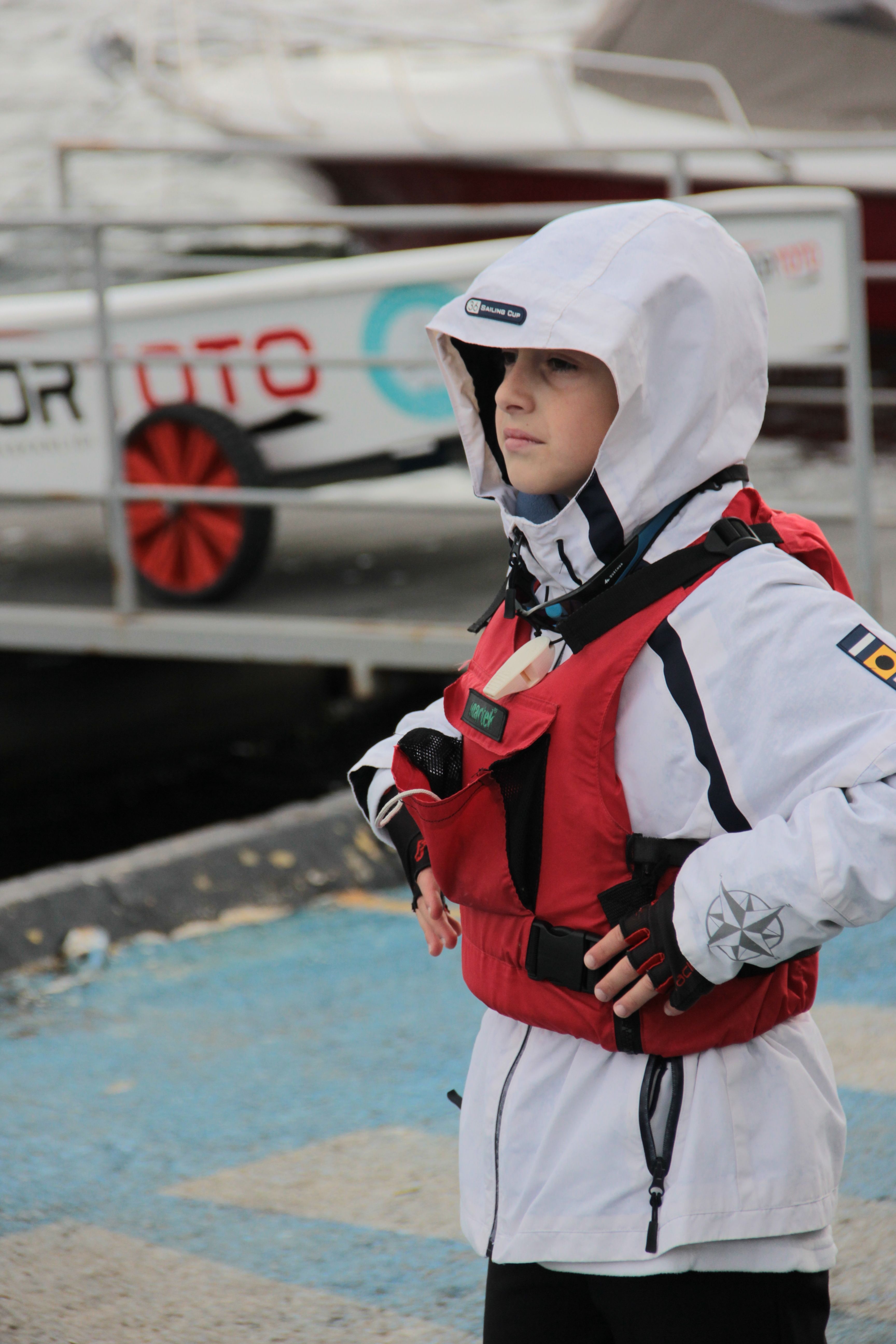 A young boy in a red life vest and white raincoat stands near a harbor, captured in a candid moment.