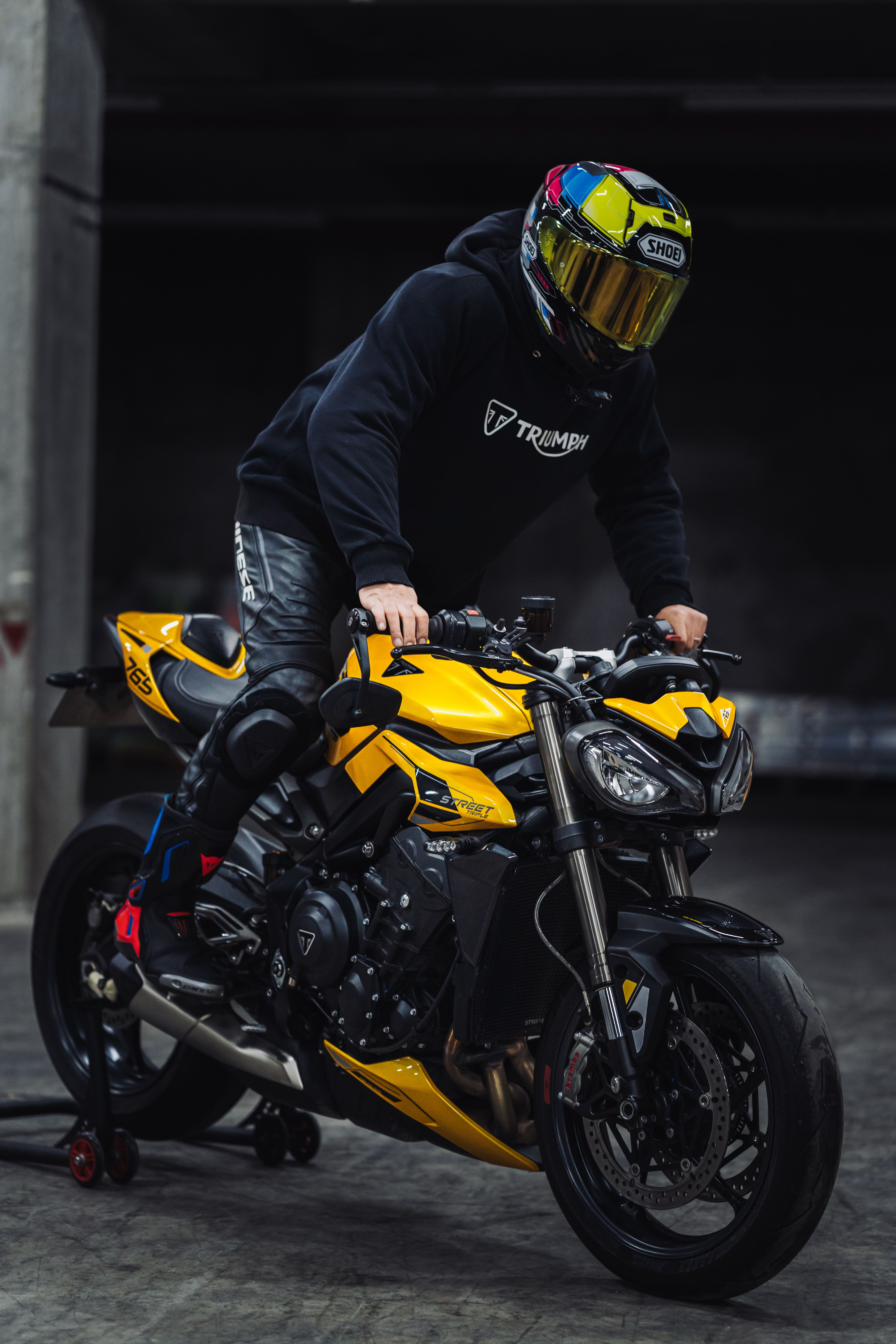 Motorcyclist with colorful helmet on a sleek yellow Triumph in an indoor setting.