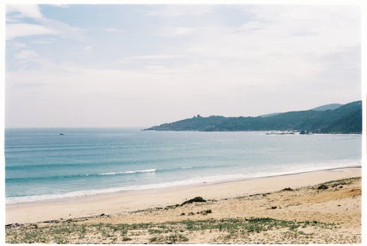 Quiet beach with gentle waves under a cloudy sky, featuring distant hills.