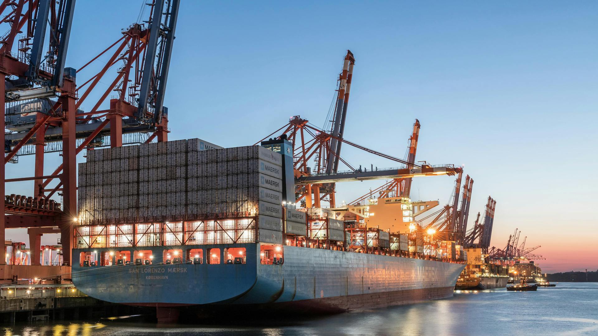 A view of a large container ship docked at Hamburg Port during twilight, showcasing industrial shipping cranes.