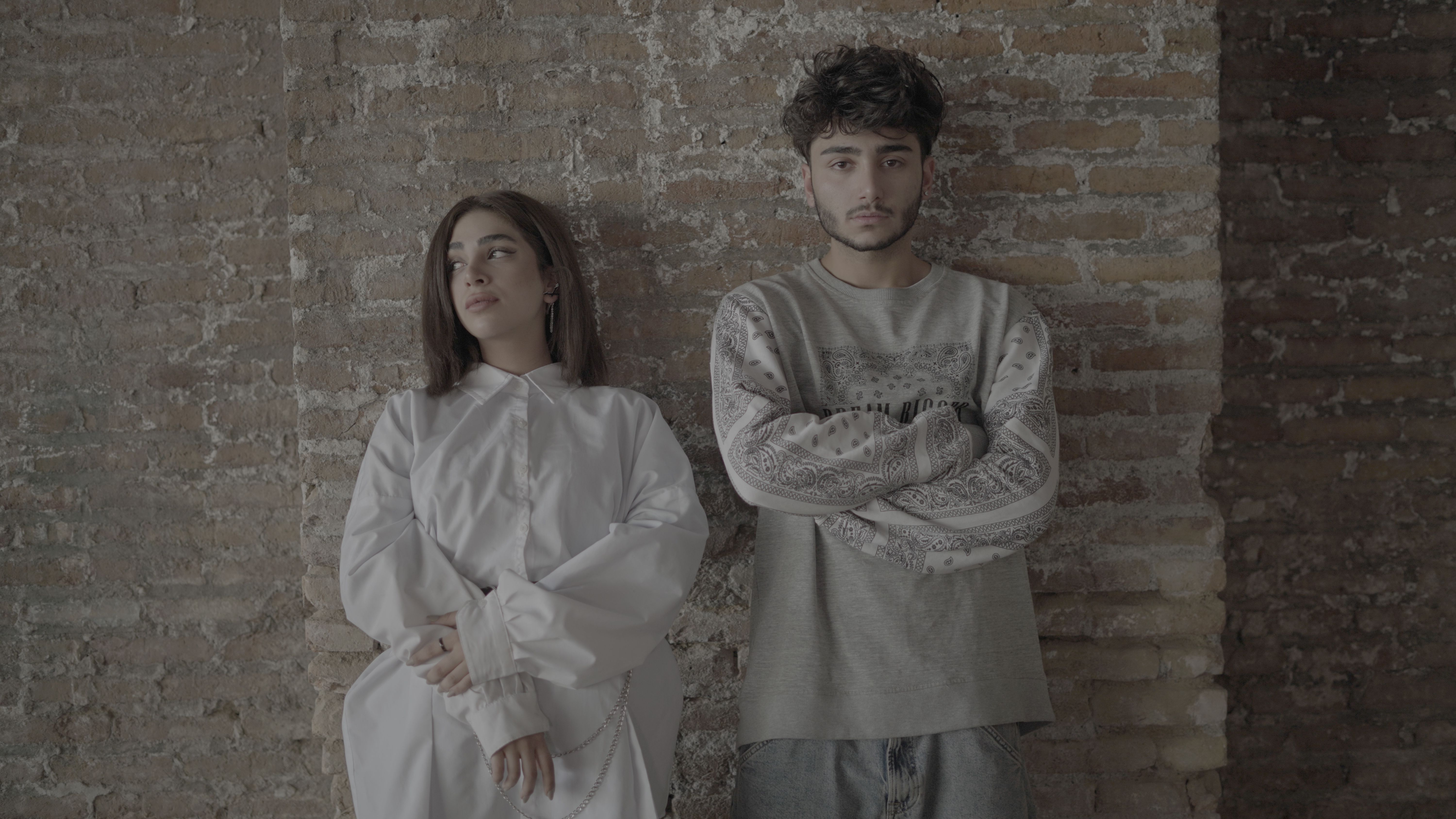 Young man and woman standing against a rustic brick wall, showcasing modern fashion and style.