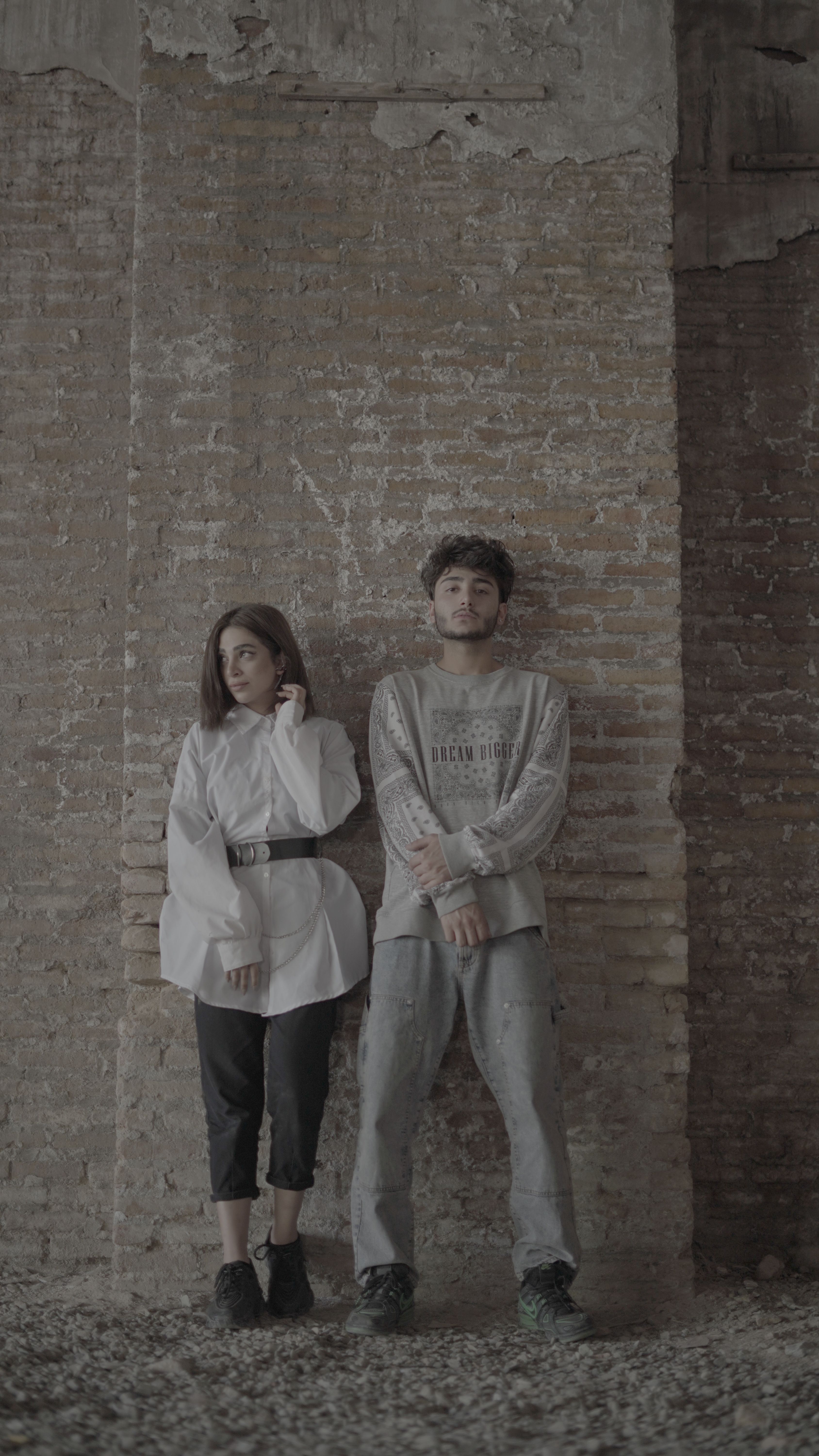 Two adults pose against a textured brick wall, showcasing modern casual fashion.