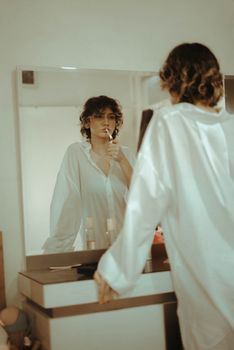 Woman in a white shirt stands thoughtfully gazing at her reflection in a mirror.