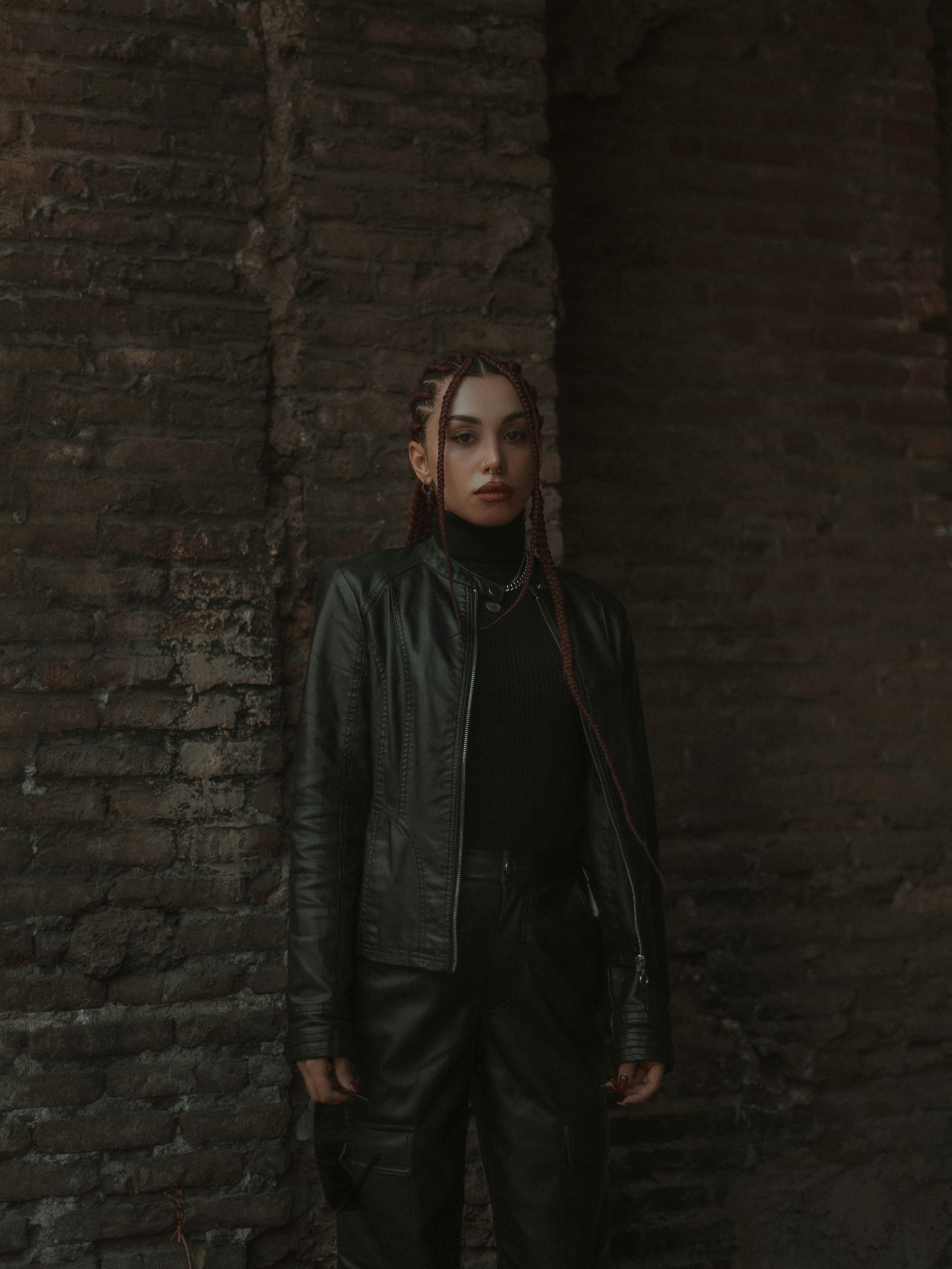 Fashionable young woman in leather jacket posing against rustic brick wall with dramatic lighting.