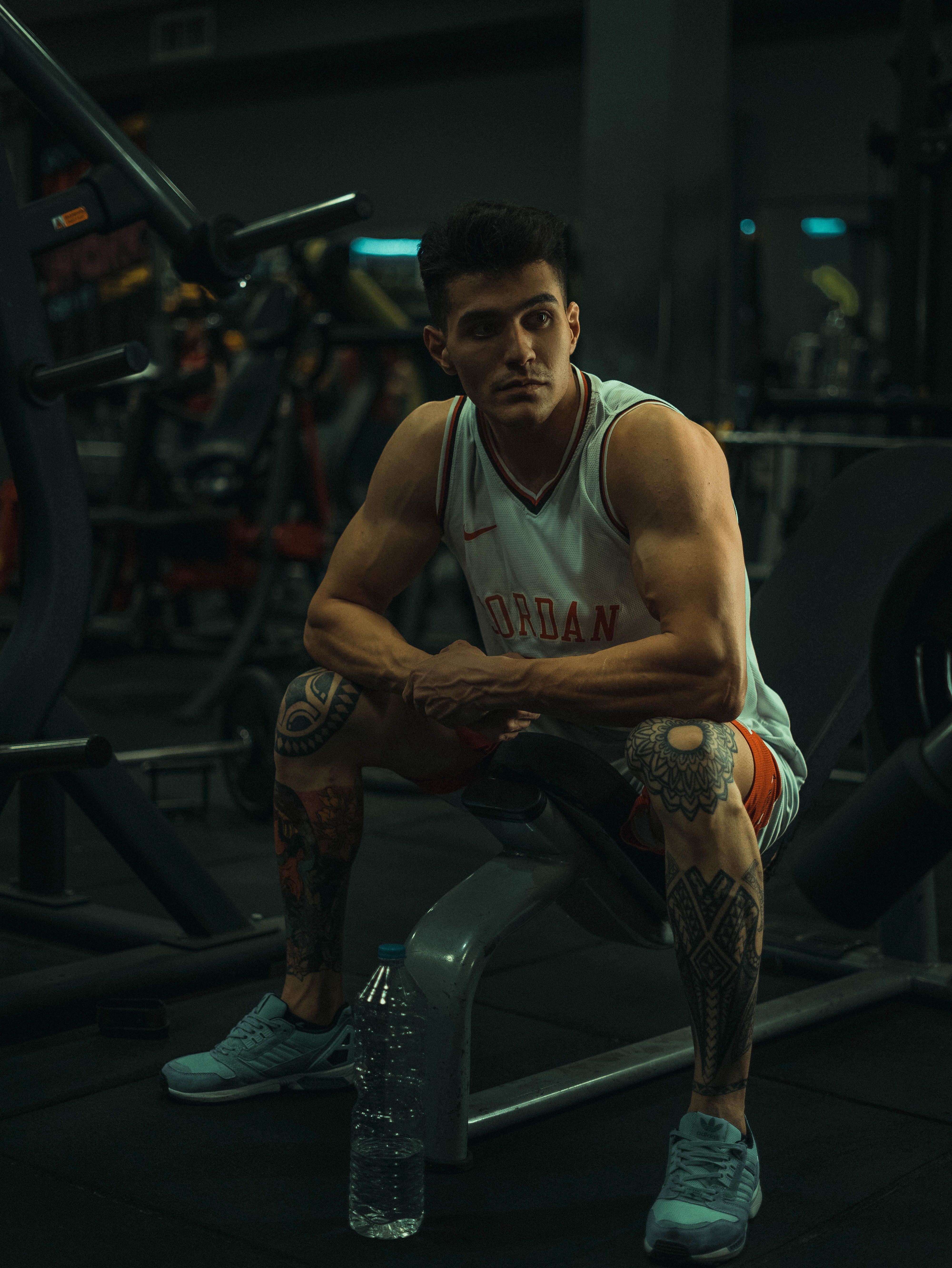 Young man with tattoos sitting on gym equipment, looking away pensively.