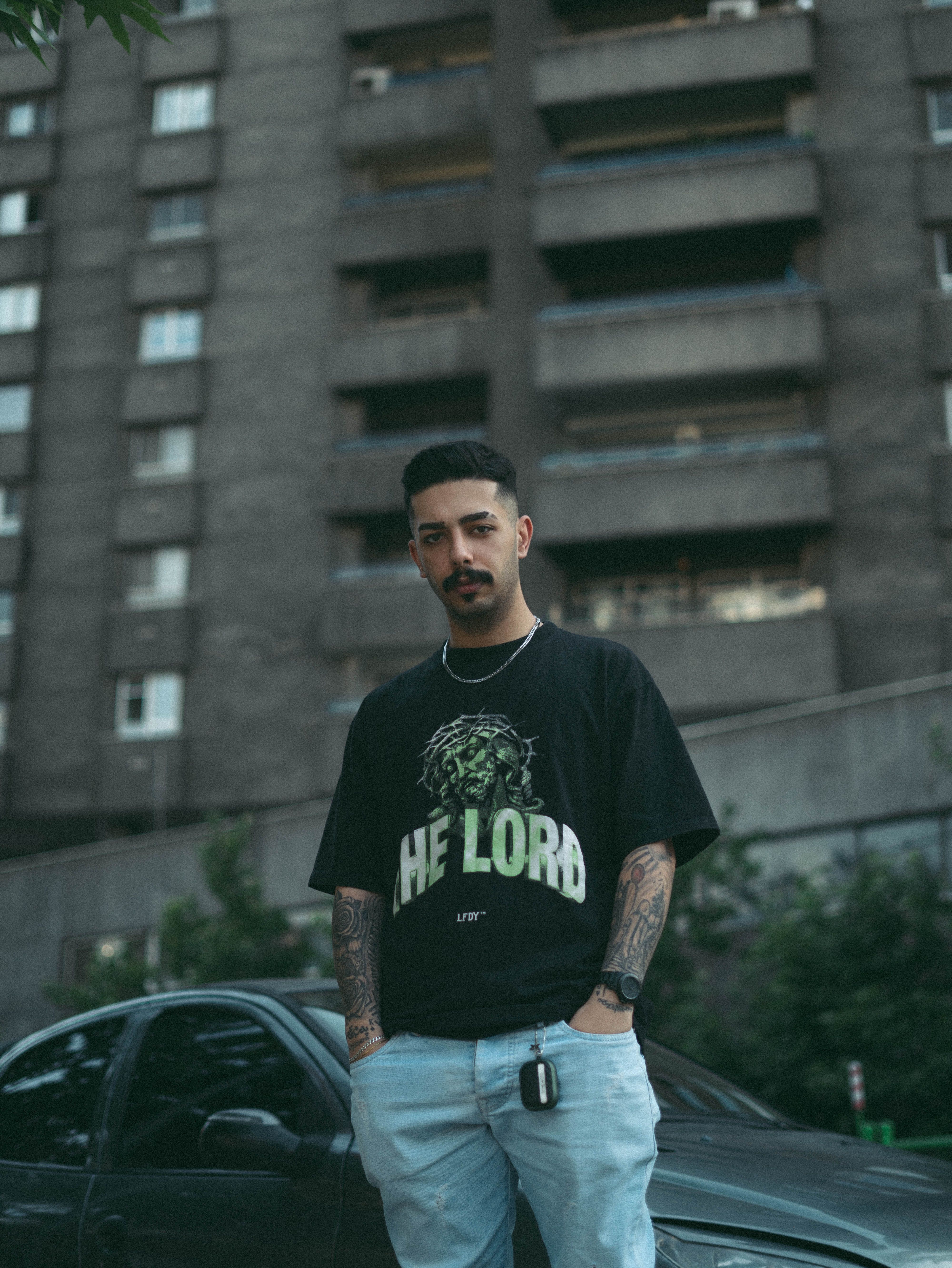 Young man with tattoos poses in front of urban apartment building.