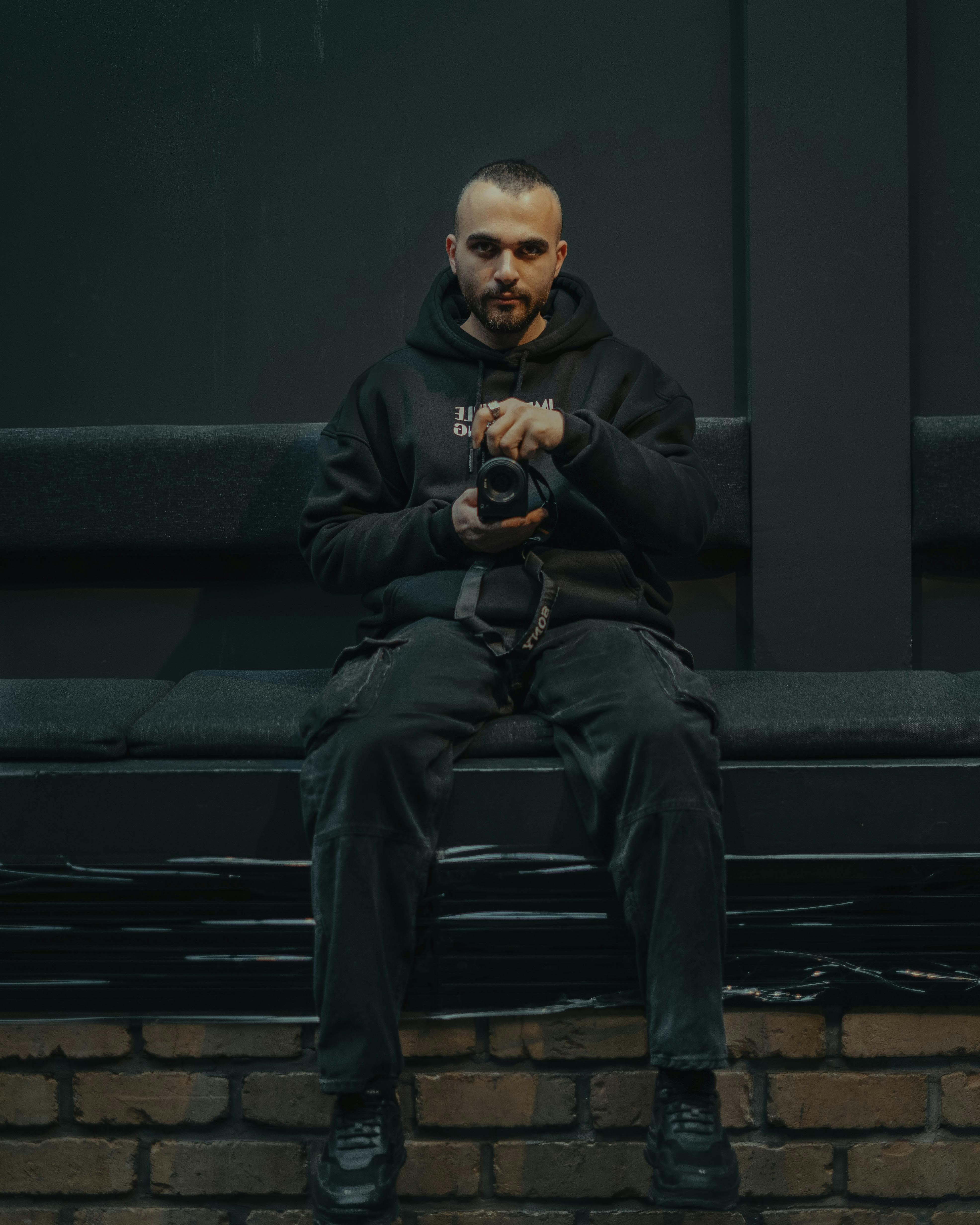 Gratis Un hombre sentado en un banco en el interior sosteniendo una cámara, vestido con ropa oscura. Foto de stock