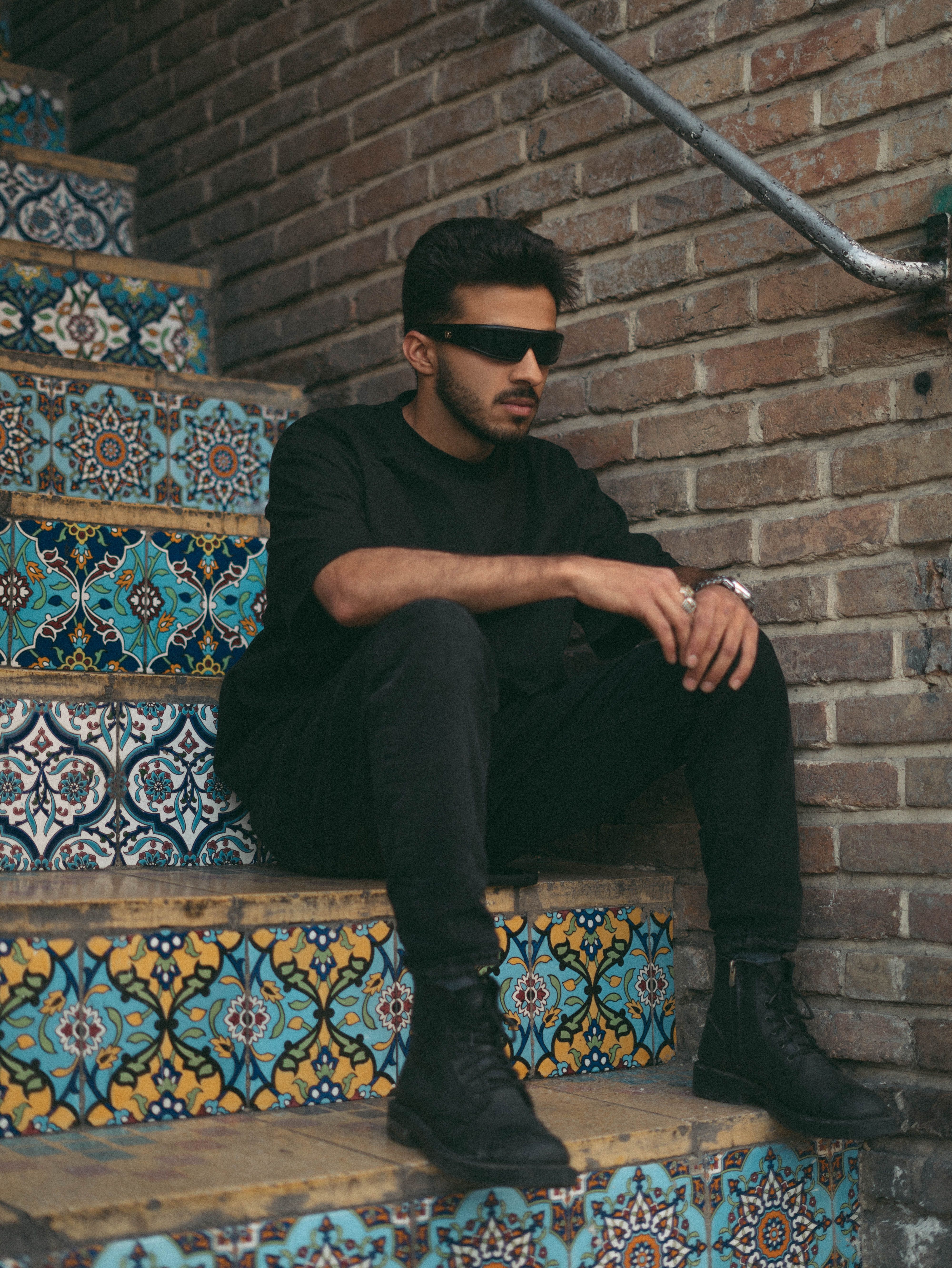 Stylish young man in sunglasses seated on vibrant tiled steps against a brick wall.