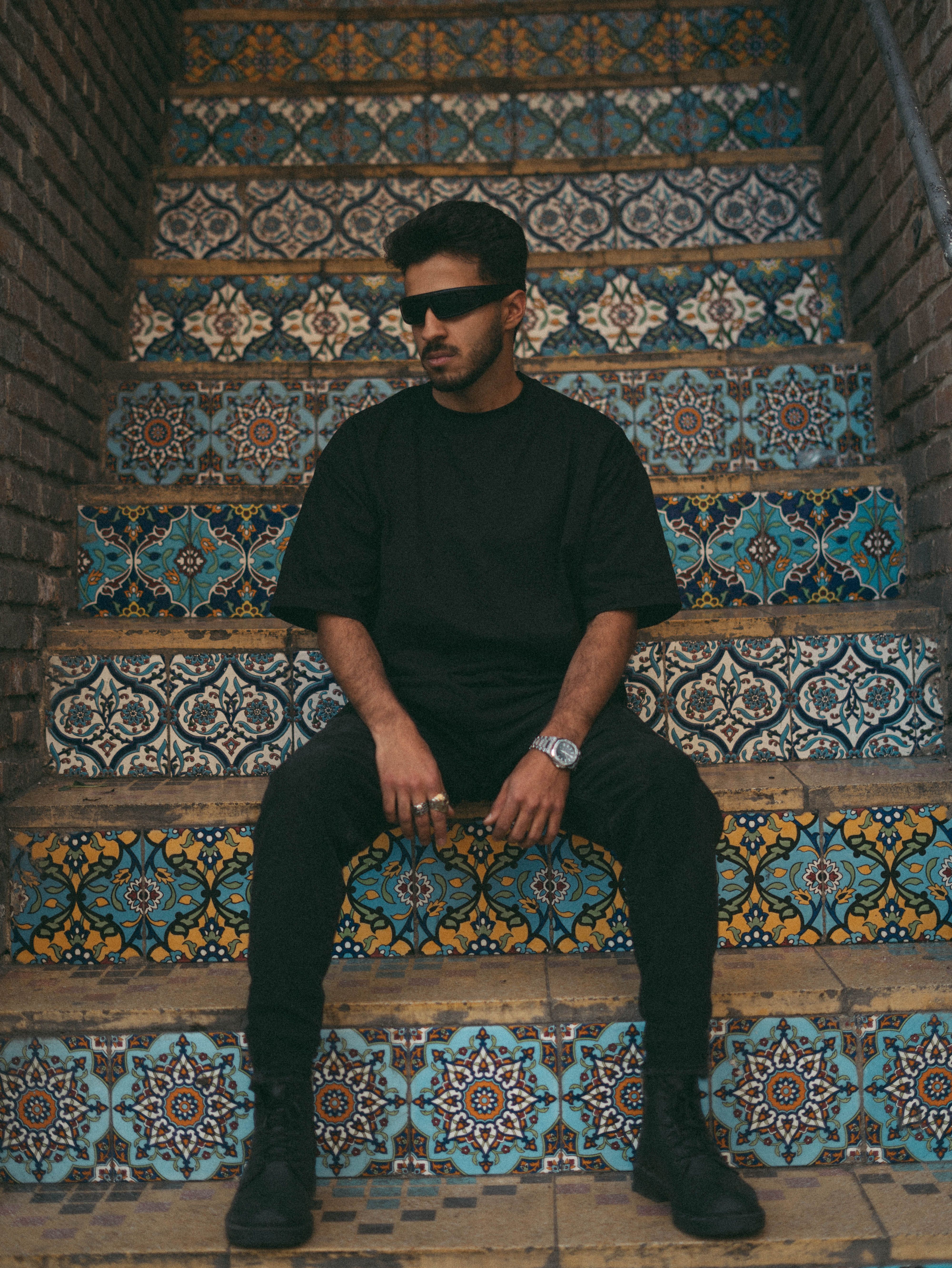 Fashionable man in black sits on colorful mosaic stairs wearing sunglasses and watch, showcasing urban style.