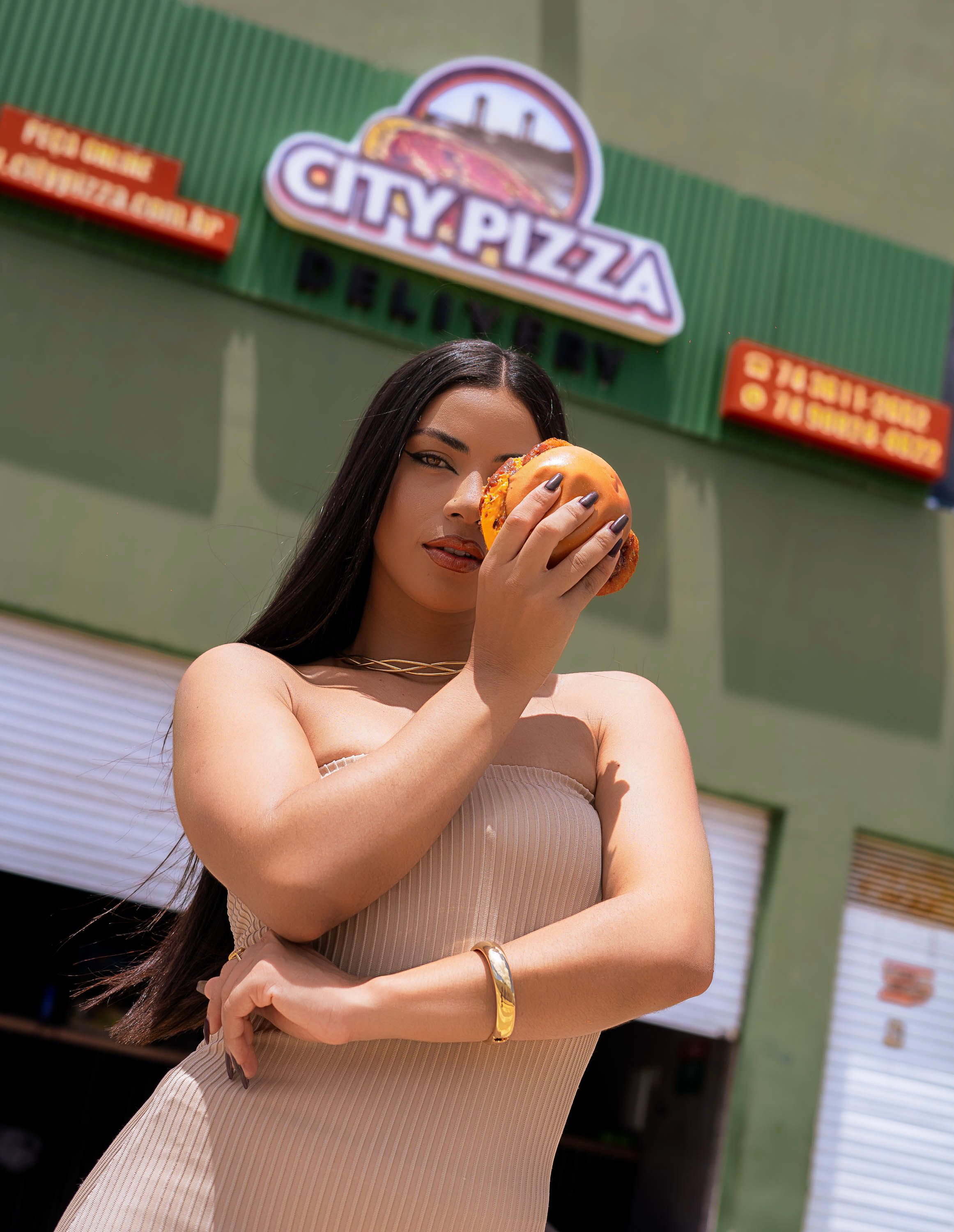 Stylish adult woman poses with a burger in front of a City Pizza sign, exuding confidence and modern lifestyle.