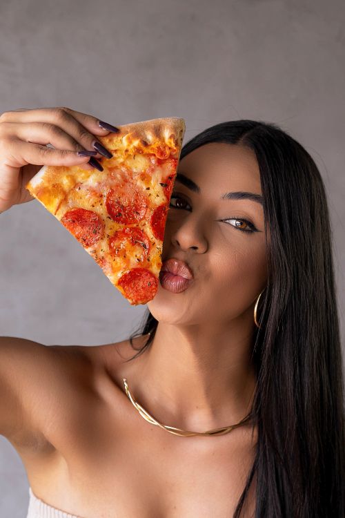 Free Portrait of a woman playfully holding a slice of pepperoni pizza near her face. Stock Photo