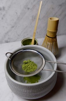 Matcha tea being sifted with a strainer over a bowl, featuring a bamboo whisk and scoop on a marble table.