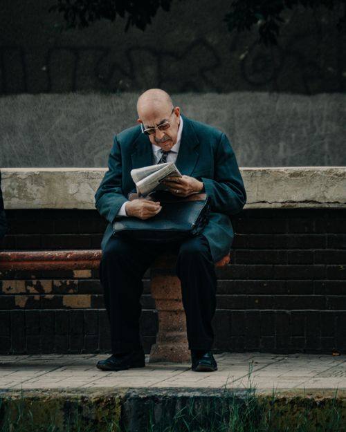 Free An elderly man in a suit sits on a bench, engrossed in a newspaper. Urban setting. Stock Photo