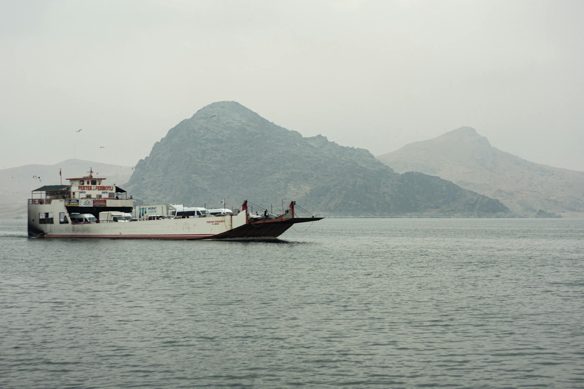A car ferry traverses calm ocean waters against a mountainous backdrop, under a cloudy sky.