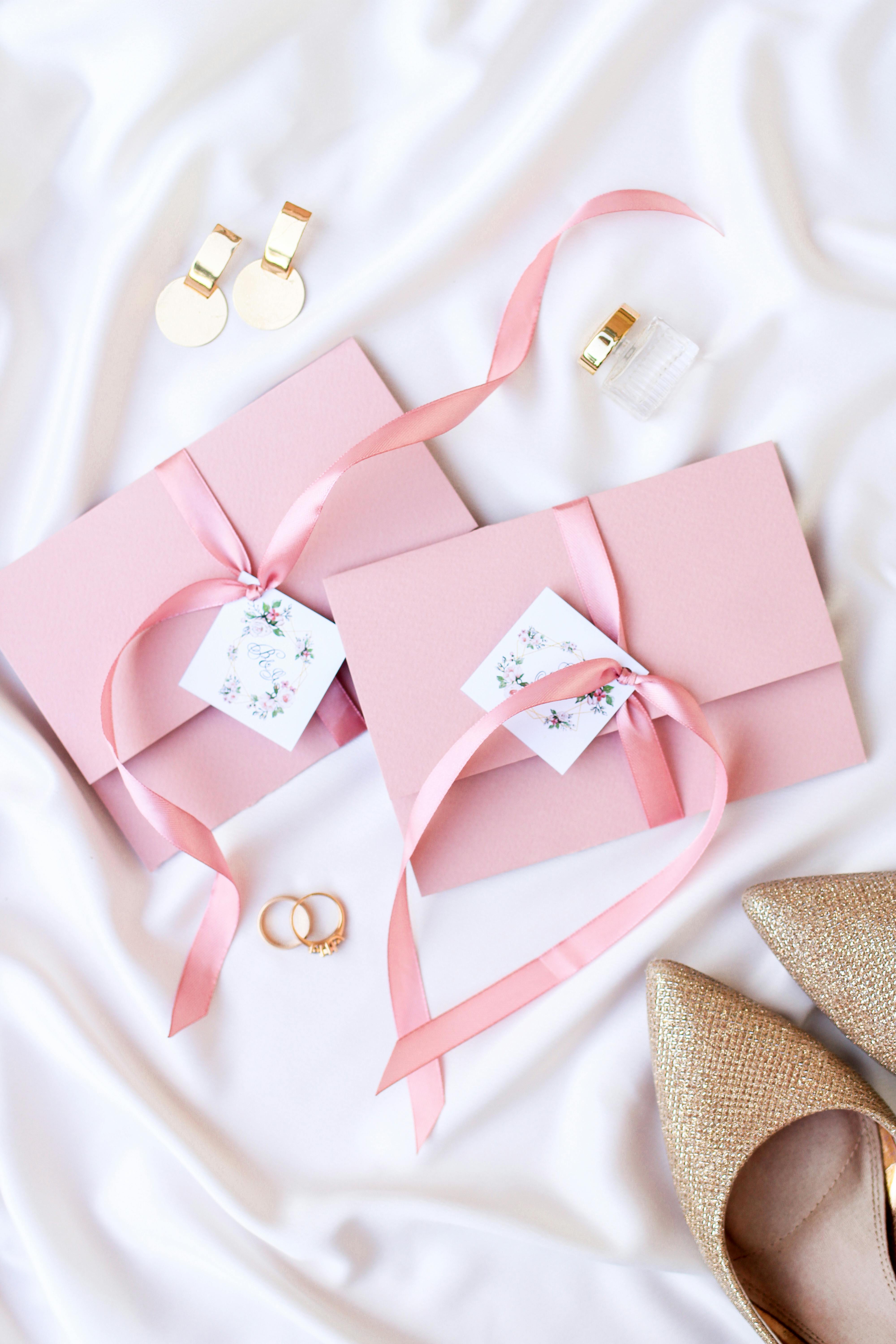 Flat lay of stylish wedding invitation with pink envelopes and accessories on satin fabric.