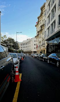 A city street lined with parked cars and classic building architecture under a clear blue sky.