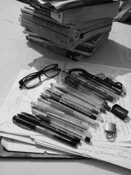 Black and white study desk with books, pens, glasses, and notes.