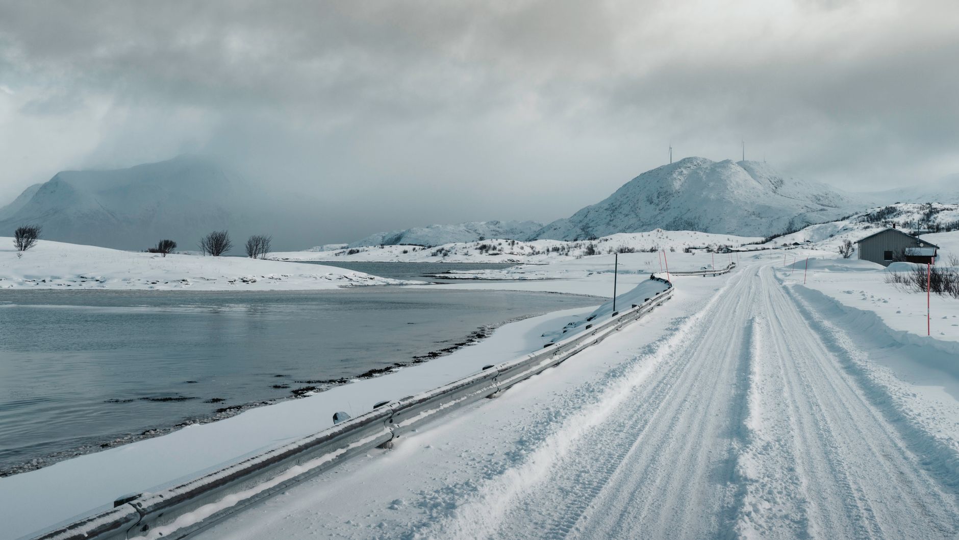 Snow-covered road in Norway with mountains and a lake under cloudy skies.