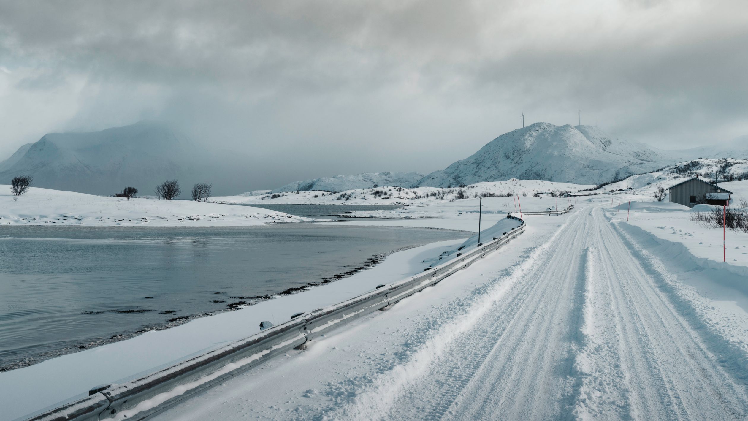 Snow-covered road in Norway with mountains and a lake under cloudy skies.
