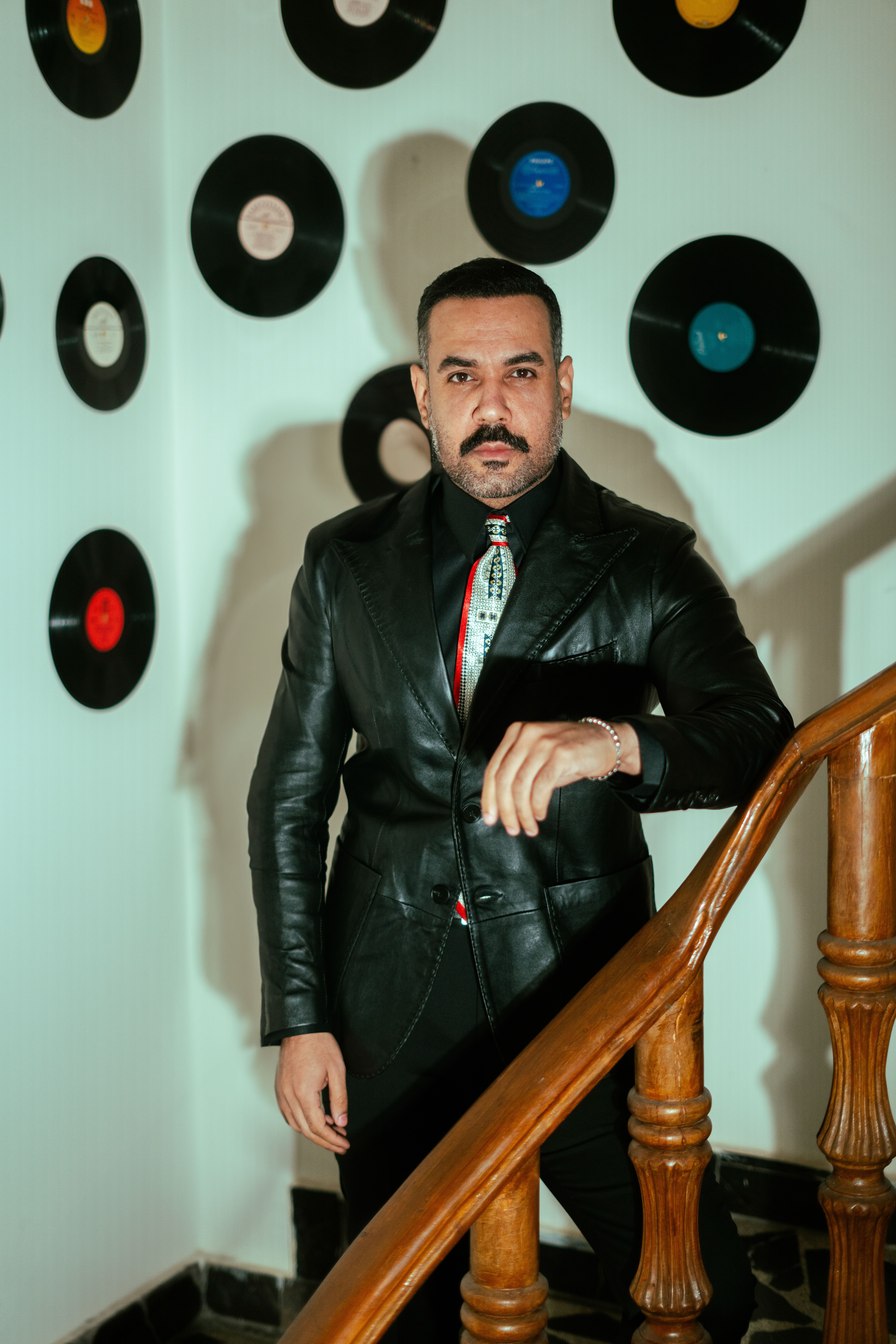 Man in a leather jacket posing on stairs, surrounded by vinyl records in Baghdad, Iraq.