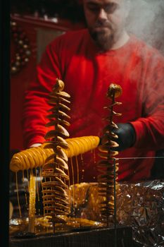 A street vendor in a red sweater preparing cheese-covered spiral potato skewers.