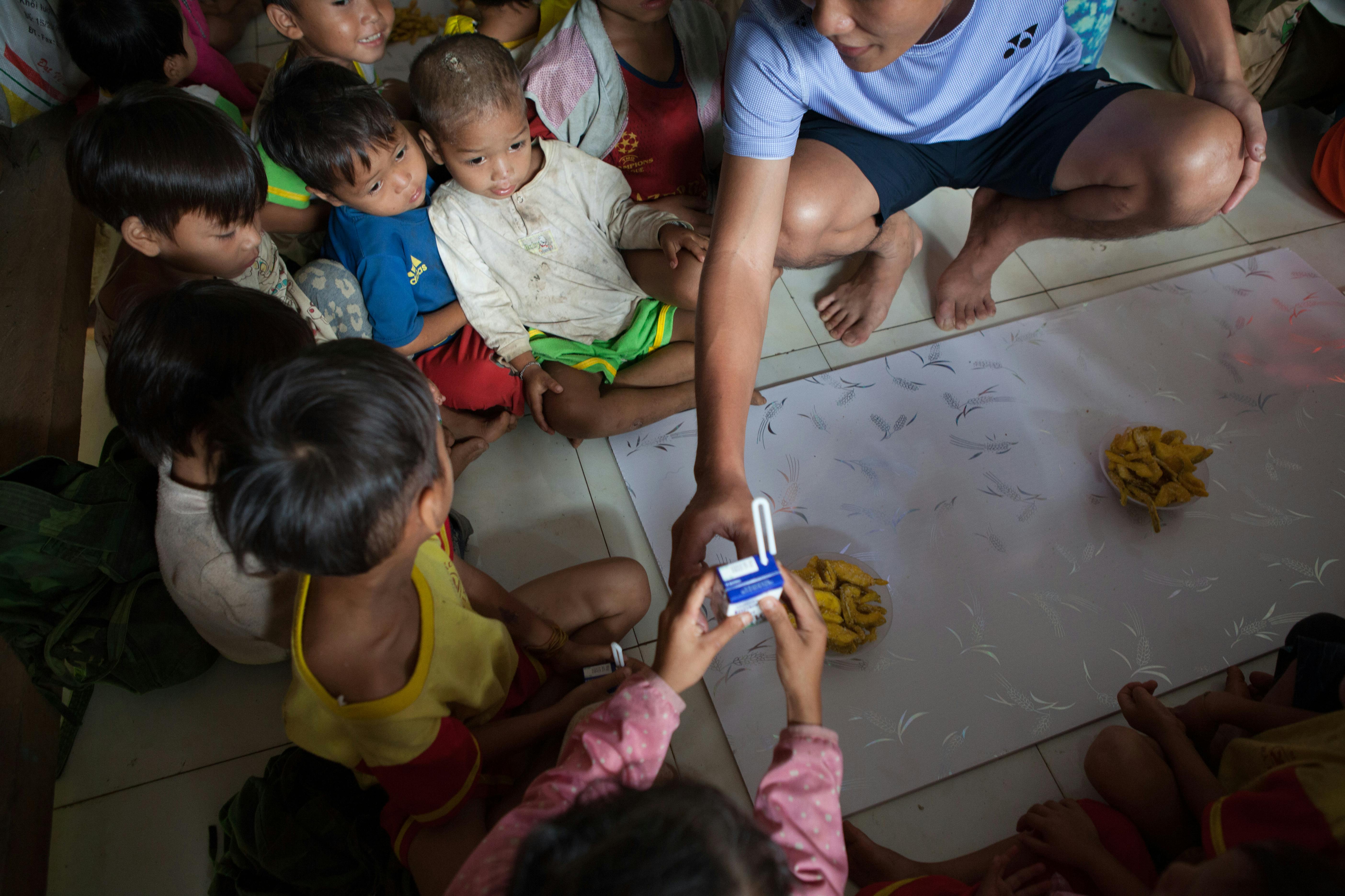 Children gather on the floor as an adult distributes milk cartons in a community setting.
