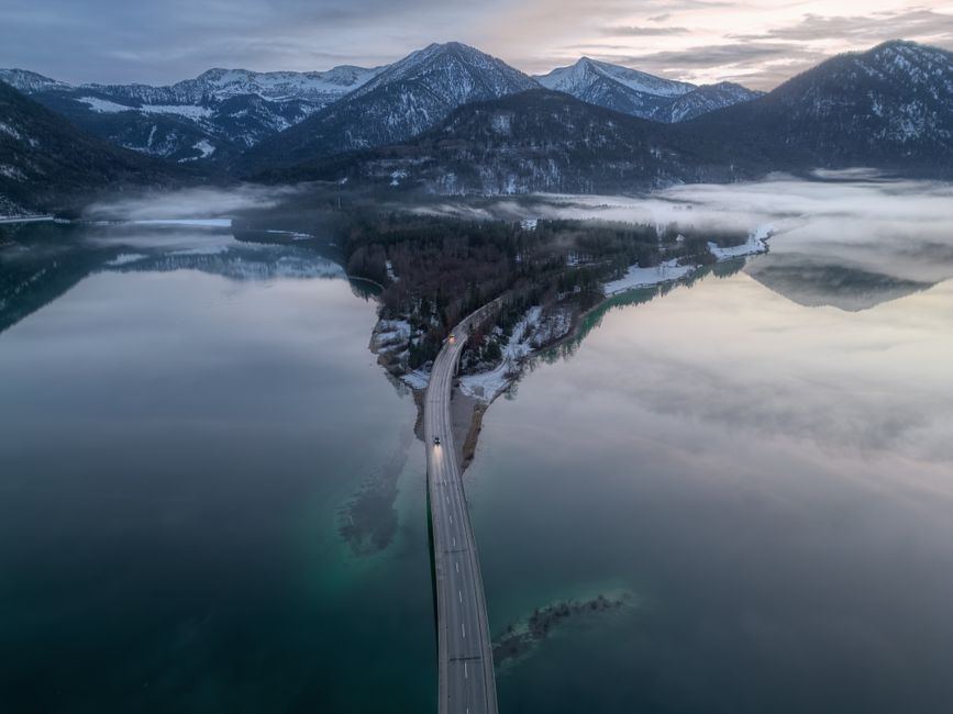 Aerial view of a scenic Bavarian landscape with a road crossing a misty lake amid snow-covered mountains.