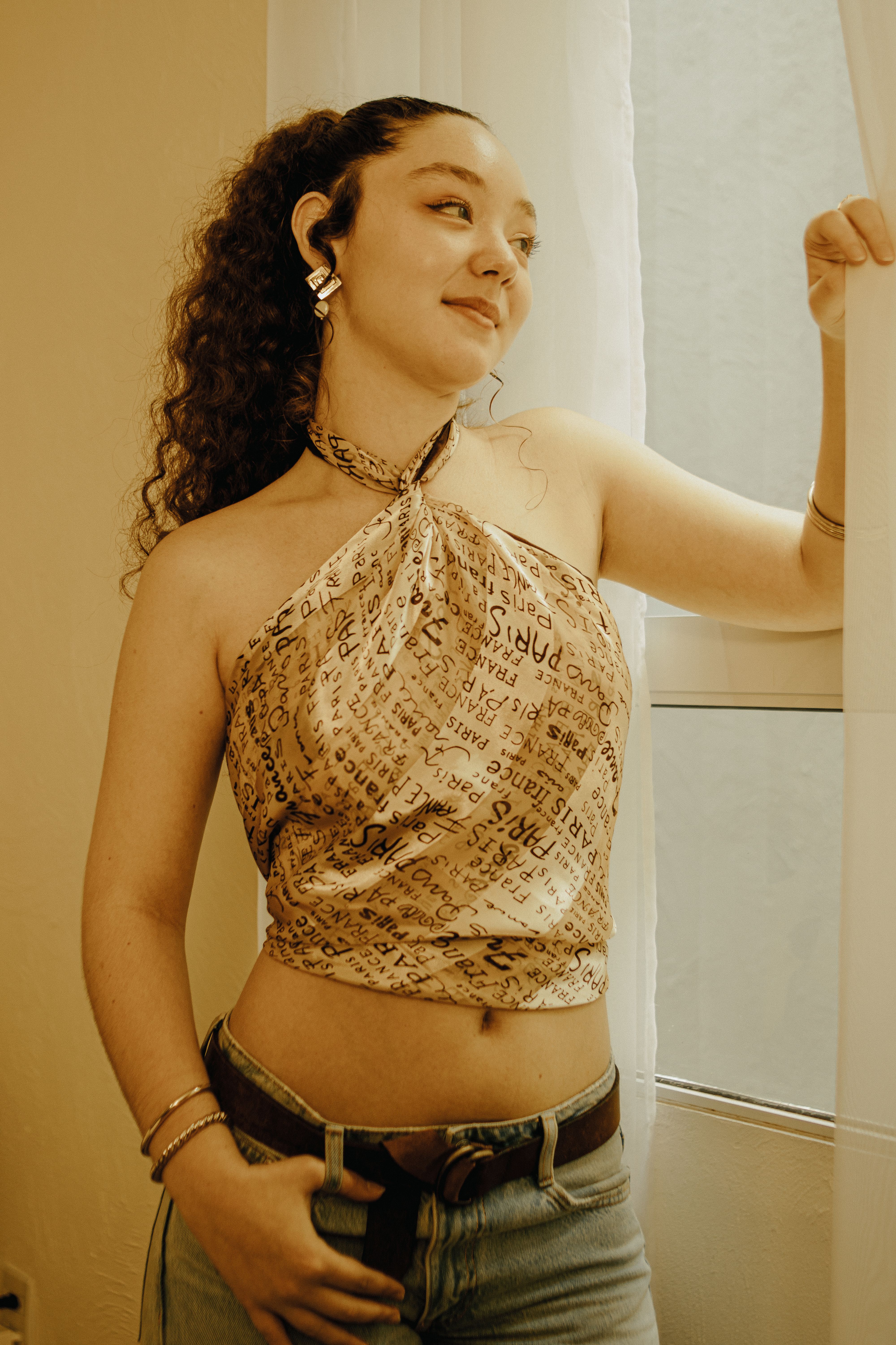 Young woman with curly hair in stylish top posing confidently indoors by a window.