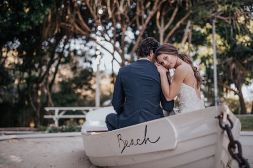 Man Wearing Blue Suit Jacket and Woman Wearing White Wedding Gown Sitting on White Canoe Boat
