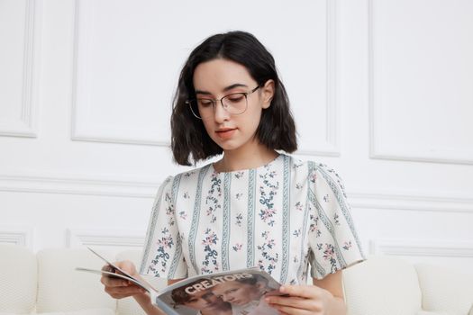 A young woman with glasses reads a magazine in a bright, minimalistic room, showcasing relaxed lifestyle and fashion.