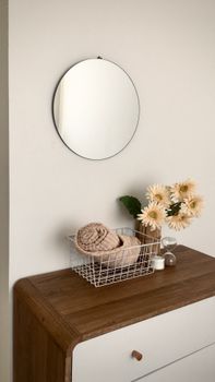 A minimalist home decor with a round mirror, daisies, and a wire basket on a wooden dresser.