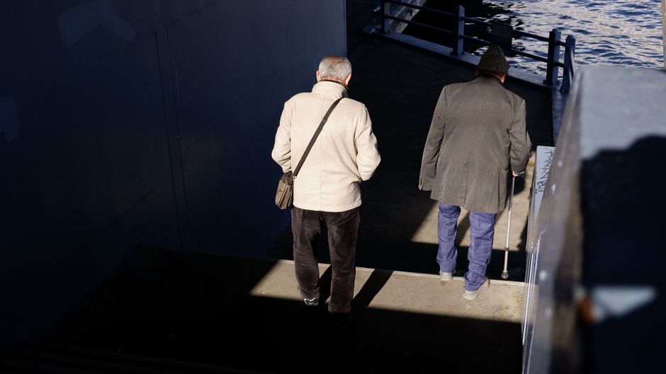 Two senior men walking down stairs outdoors, one using a cane, in bright sunlight.