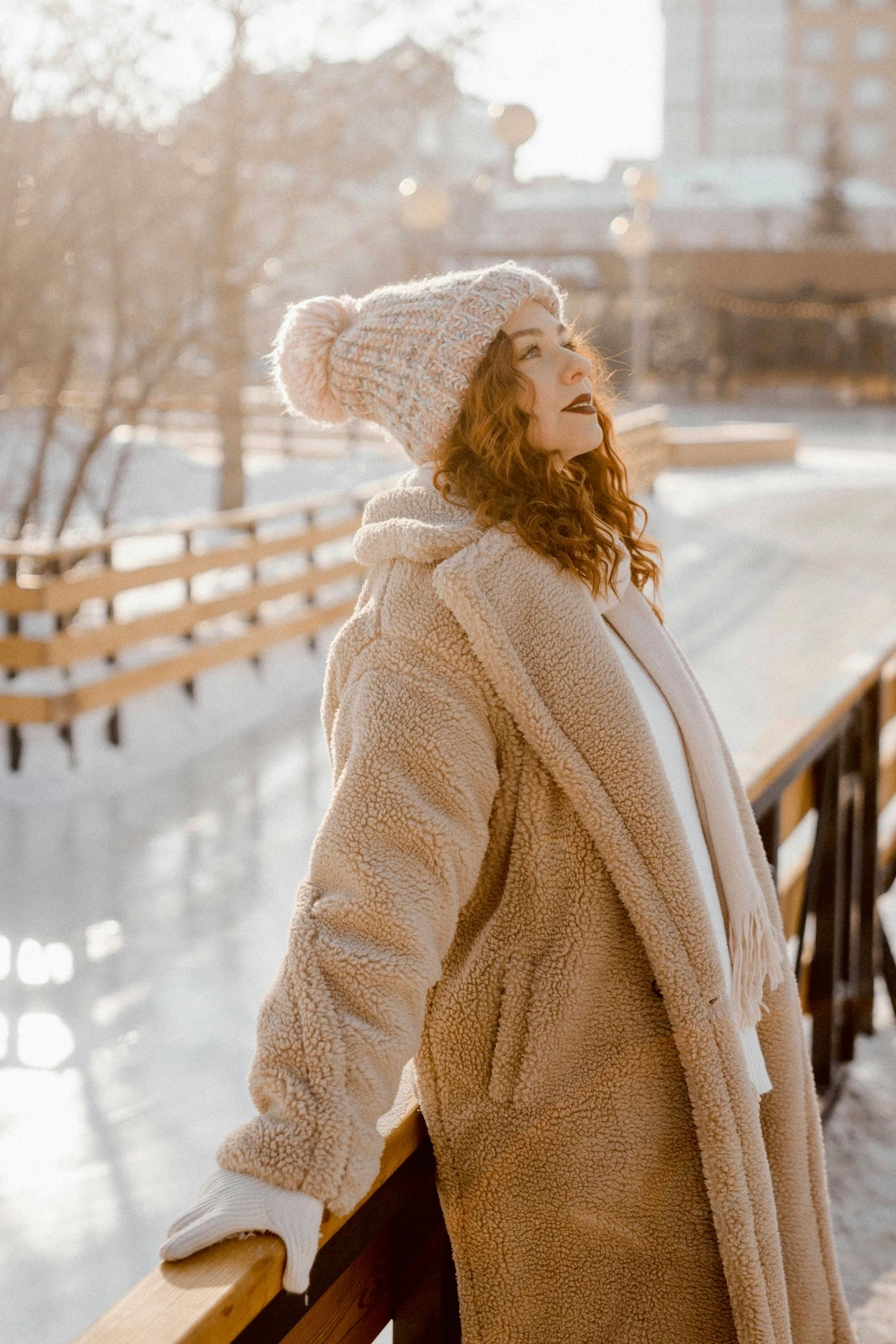 Young woman in winter attire enjoying a sunny day outside, leaning on a wooden railing.