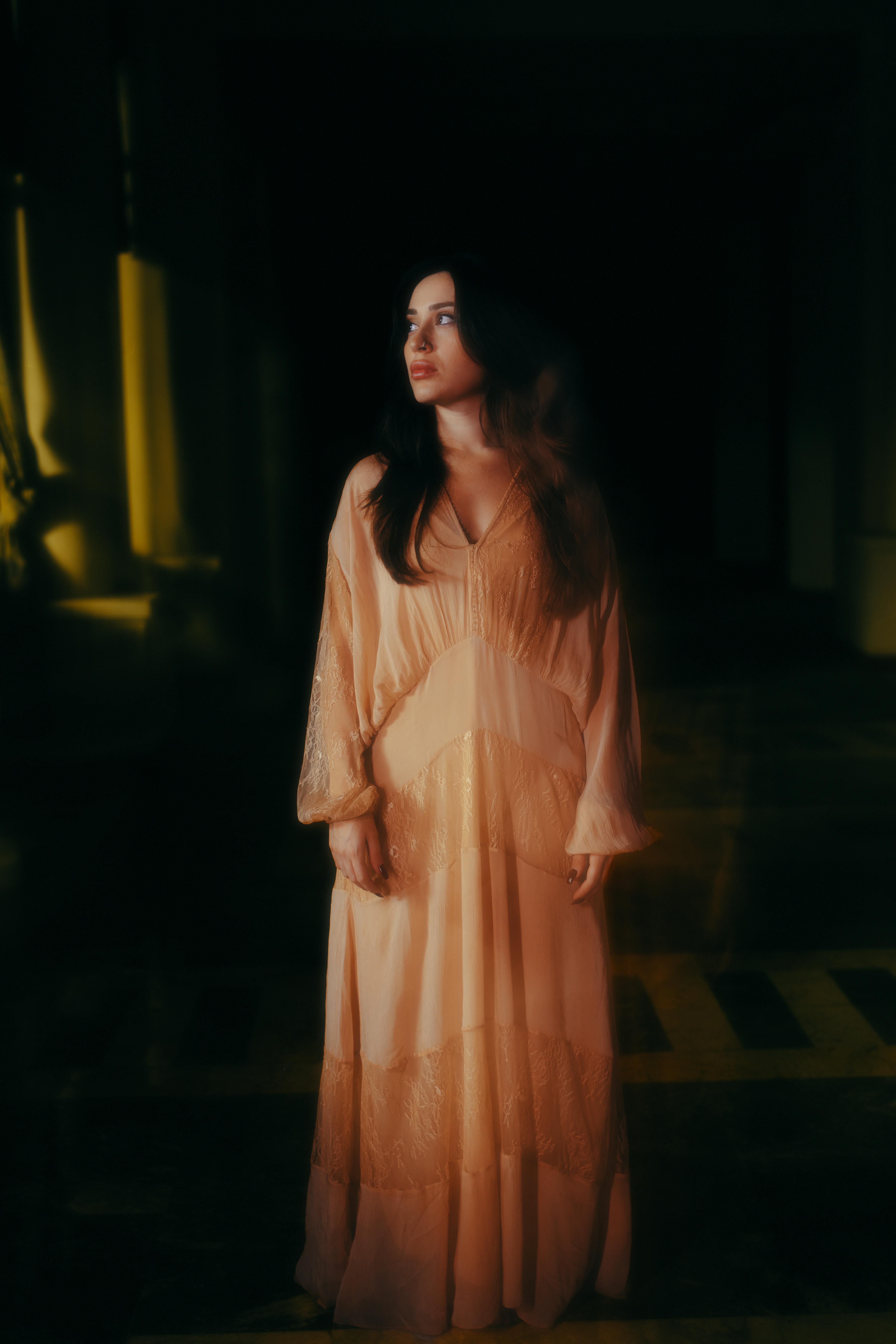 A woman in a flowing dress stands in a dimly lit indoor setting, exuding an ethereal aura.