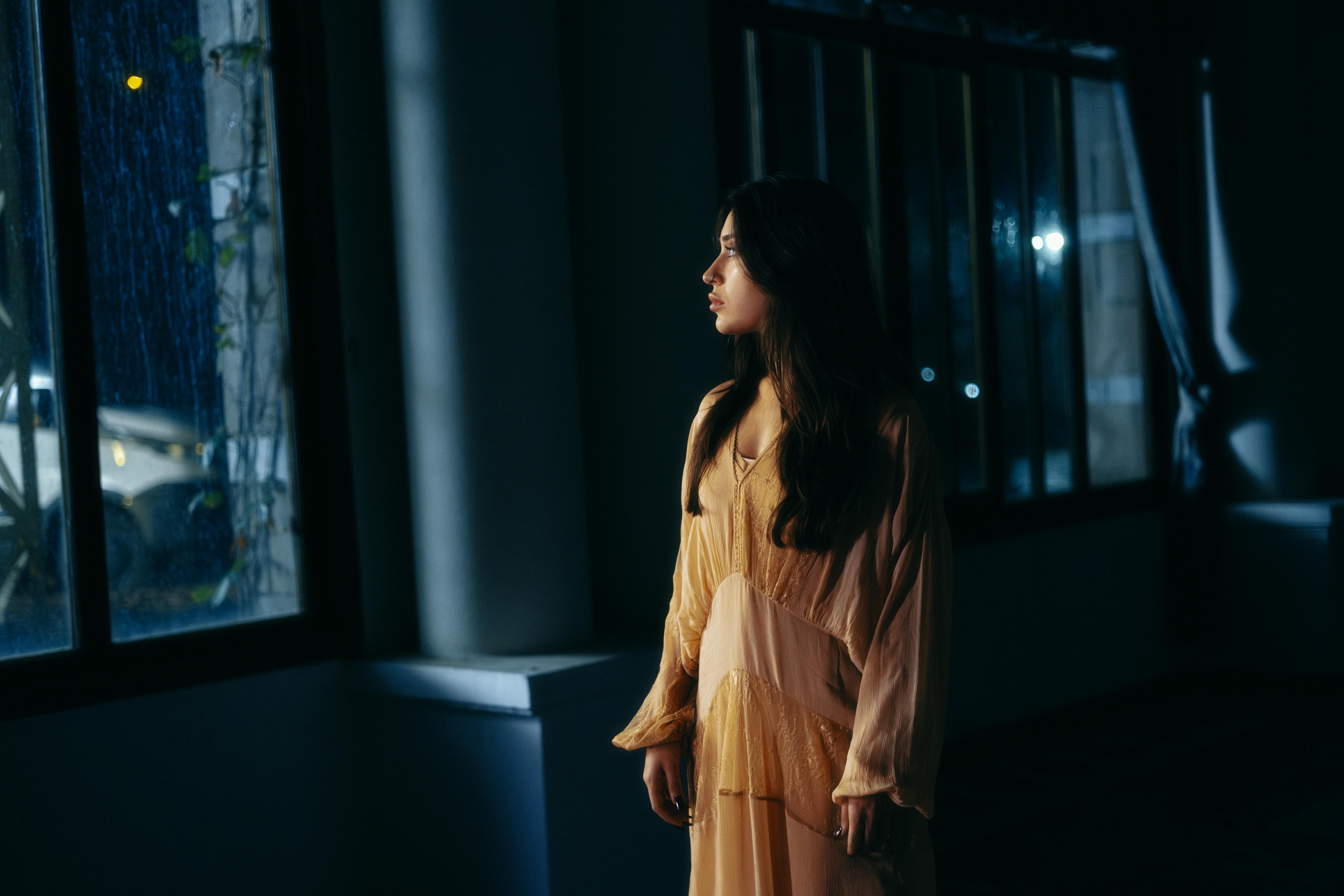 A woman in a dimly lit room looking thoughtfully out a rain-streaked window.