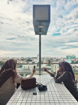 Two women capturing city views on a rooftop in Tegalsari, Indonesia, with smartphones. - Fotografer Sidoarjo