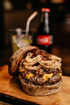 Close-up of a delicious gourmet burger topped with onion rings, cheese, and served with a bottle of cola.