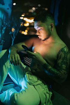 Tattooed young man engrossed in smartphone screen amid urban night lights.