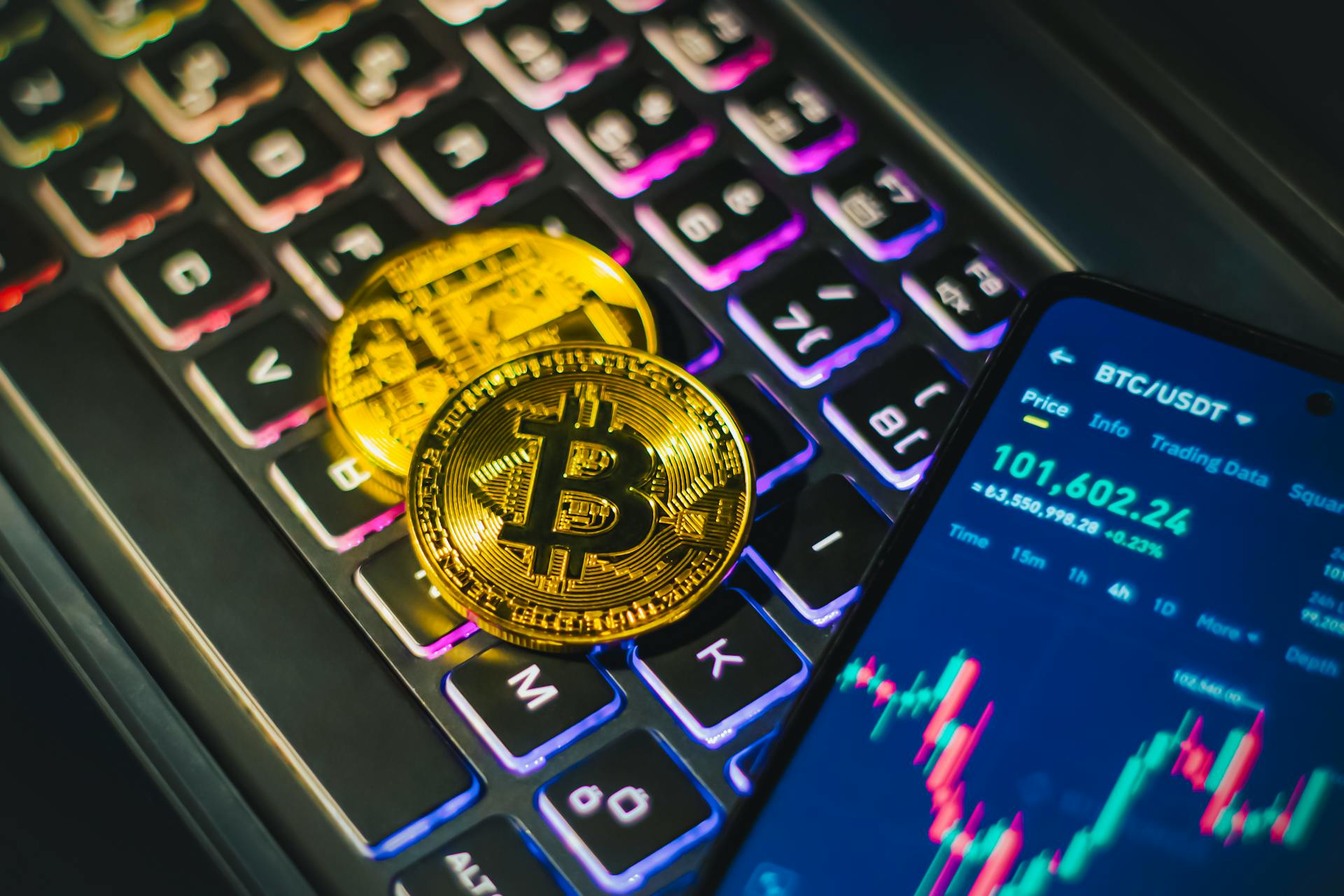 Close-up of gold Bitcoin coins on a backlit keyboard with trading app on smartphone.