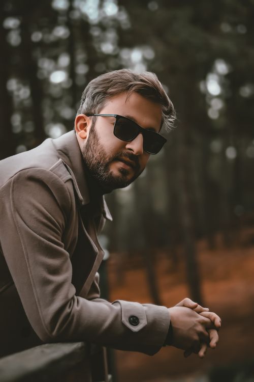 Free A fashionable man wearing sunglasses and a coat stands in a serene forest. Stock Photo