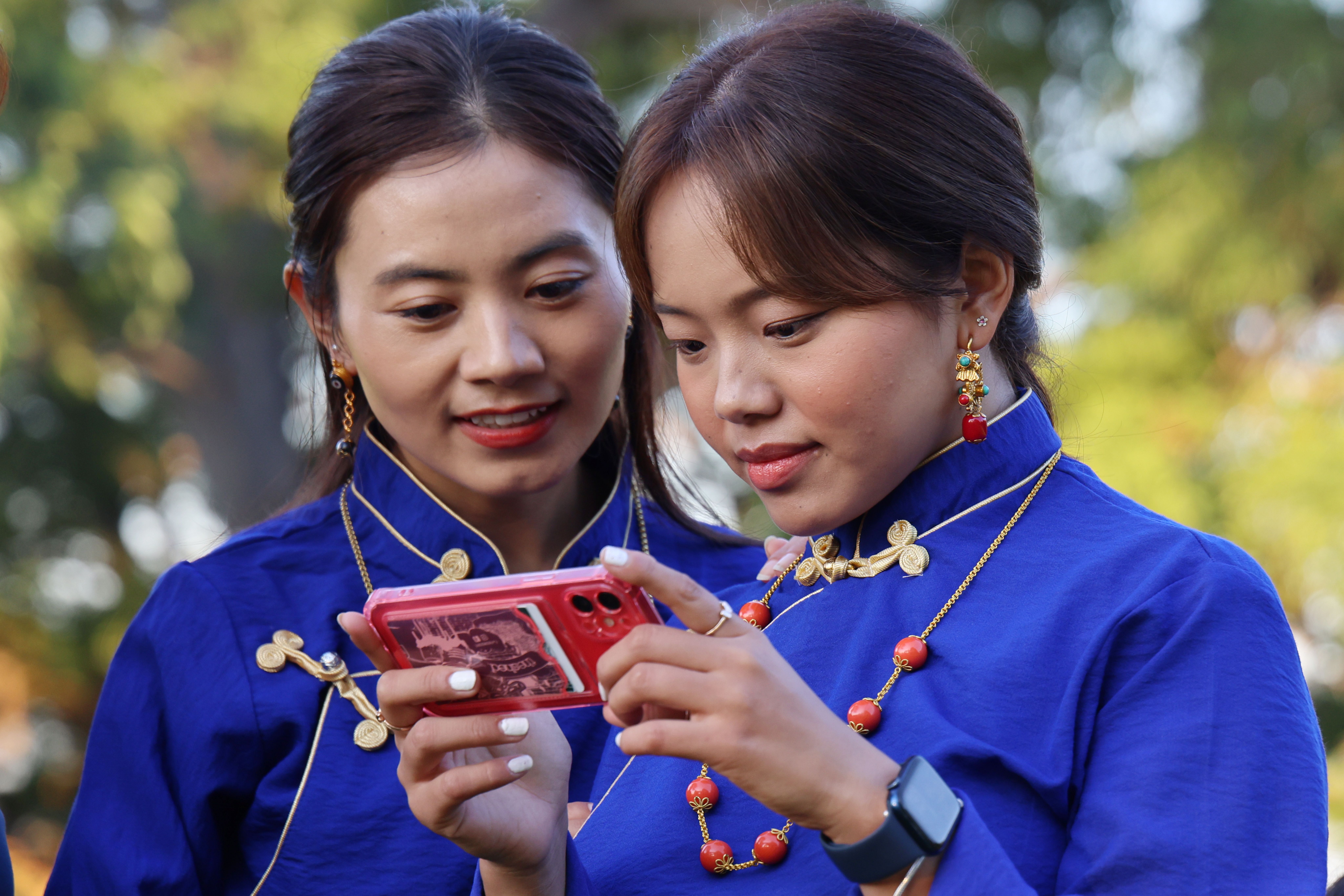 Two women in traditional blue attire using a smartphone outdoors, enjoying sunny weather.