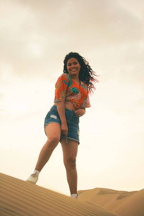 Free Smiling young woman stands confidently on a desert sand dune under a cloudy sky. Stock Photo