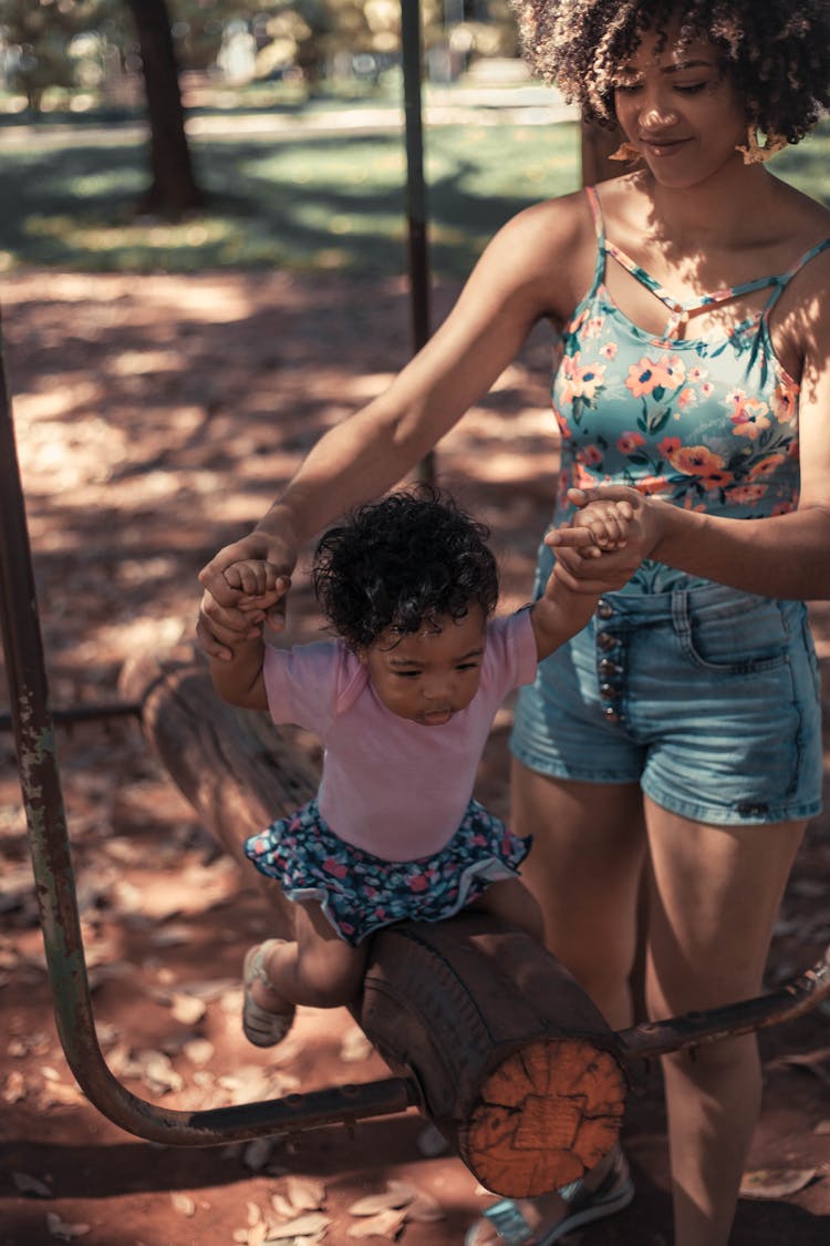 Mother Helping Her Baby Girl Sit On Seesaw