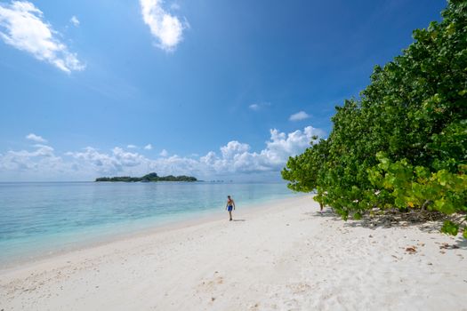 A tranquil beach scene with a person walking on white sand near lush vegetation under a bright blue sky.