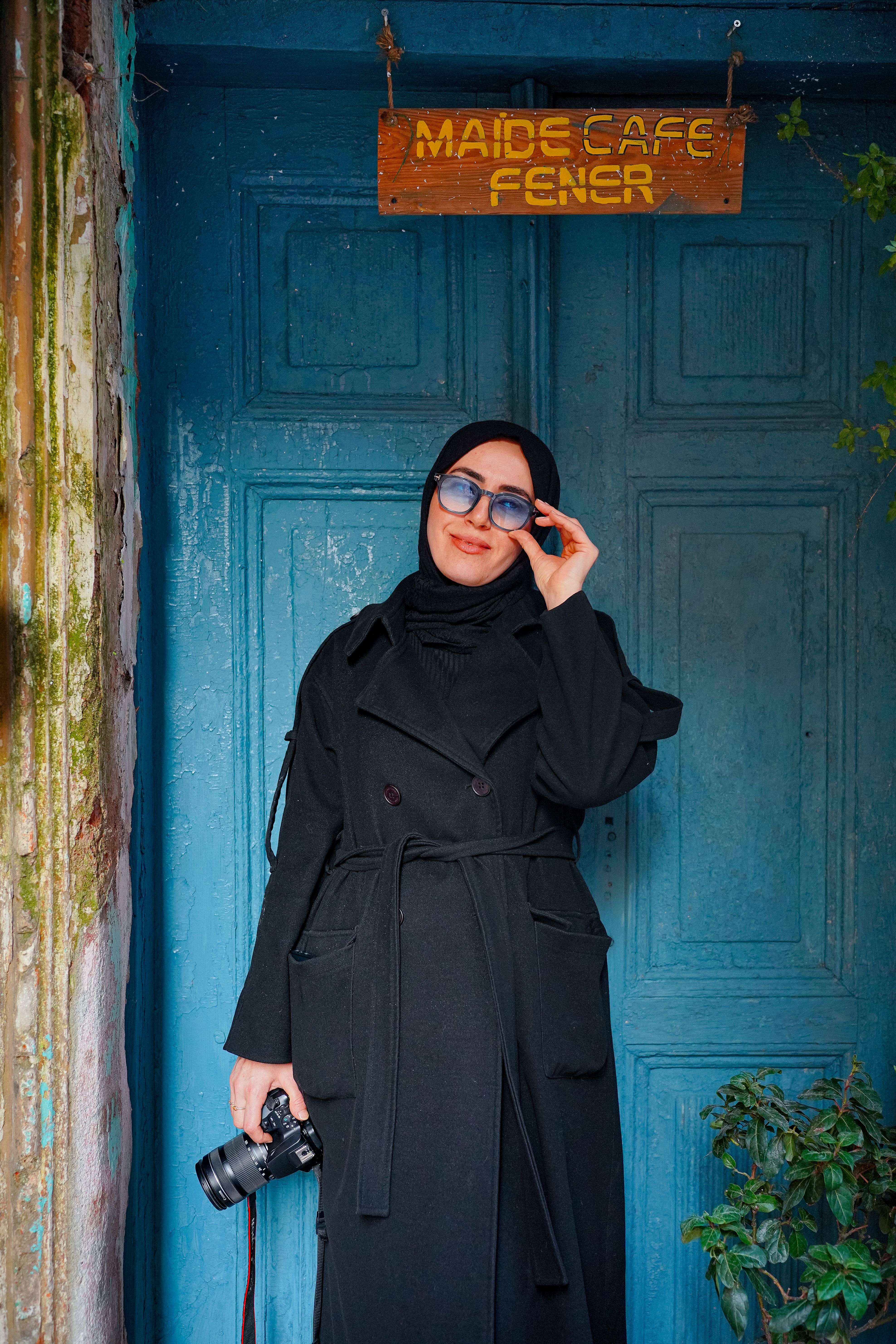Confident woman in black coat poses outdoors with camera, blue sunglasses.
