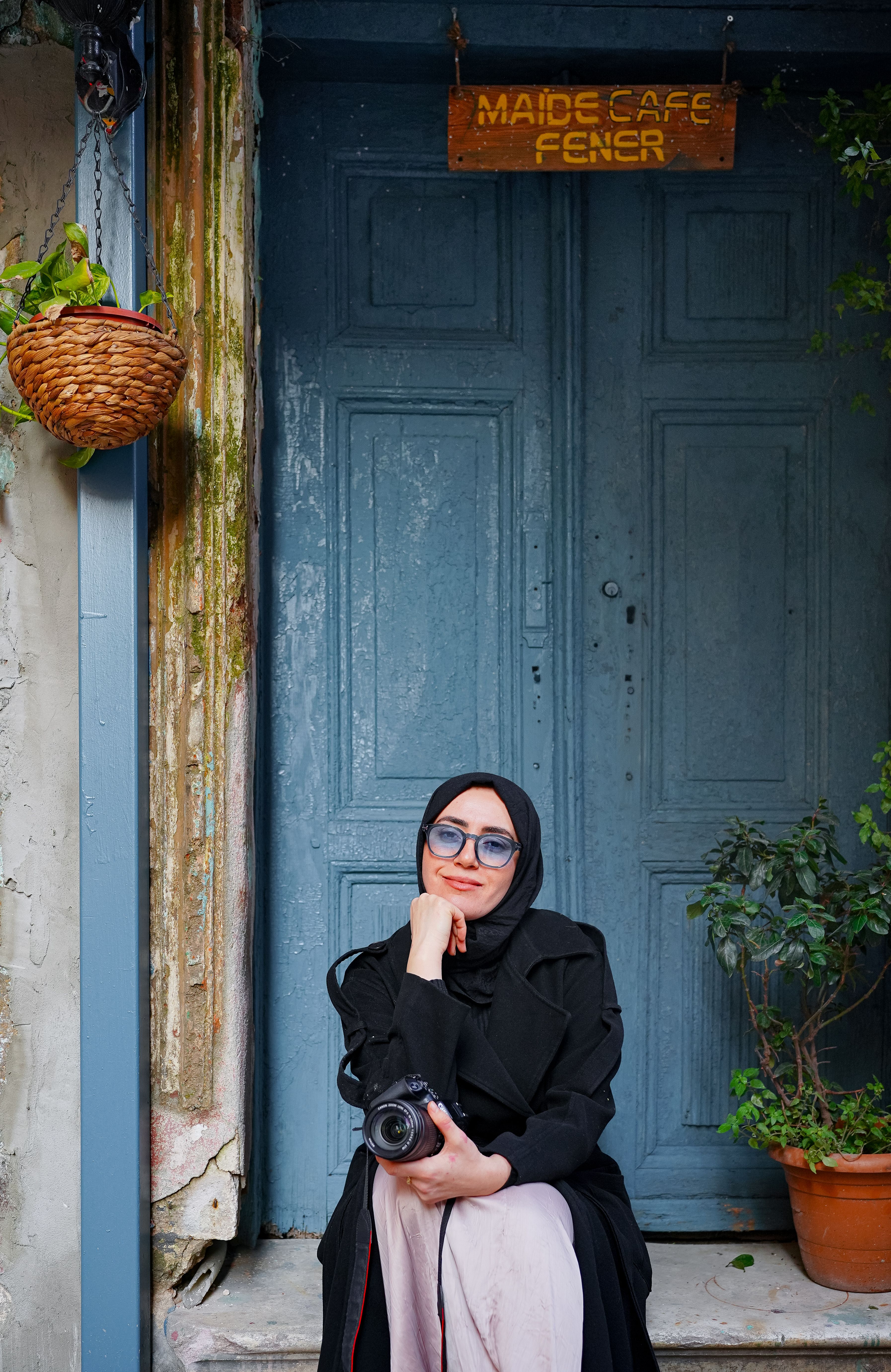 Woman in hijab sitting by a vintage door with a camera at Maide Cafe Fener.